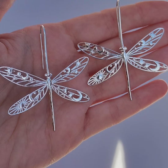 Video of a hand holding celestial damselfly earrings, showing the detailed cut‑out wings with star, moon, and sun motifs as they move in the light