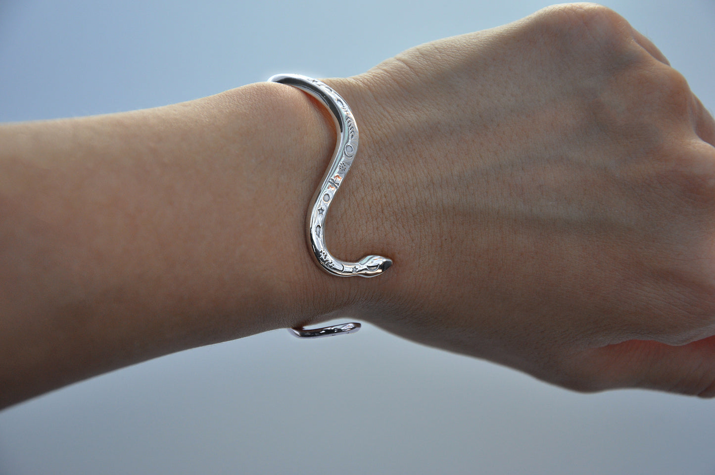 Close‑up of sterling silver snake bracelet and rings worn on the hand against a soft grey background, highlighting polished texture and engraved details.