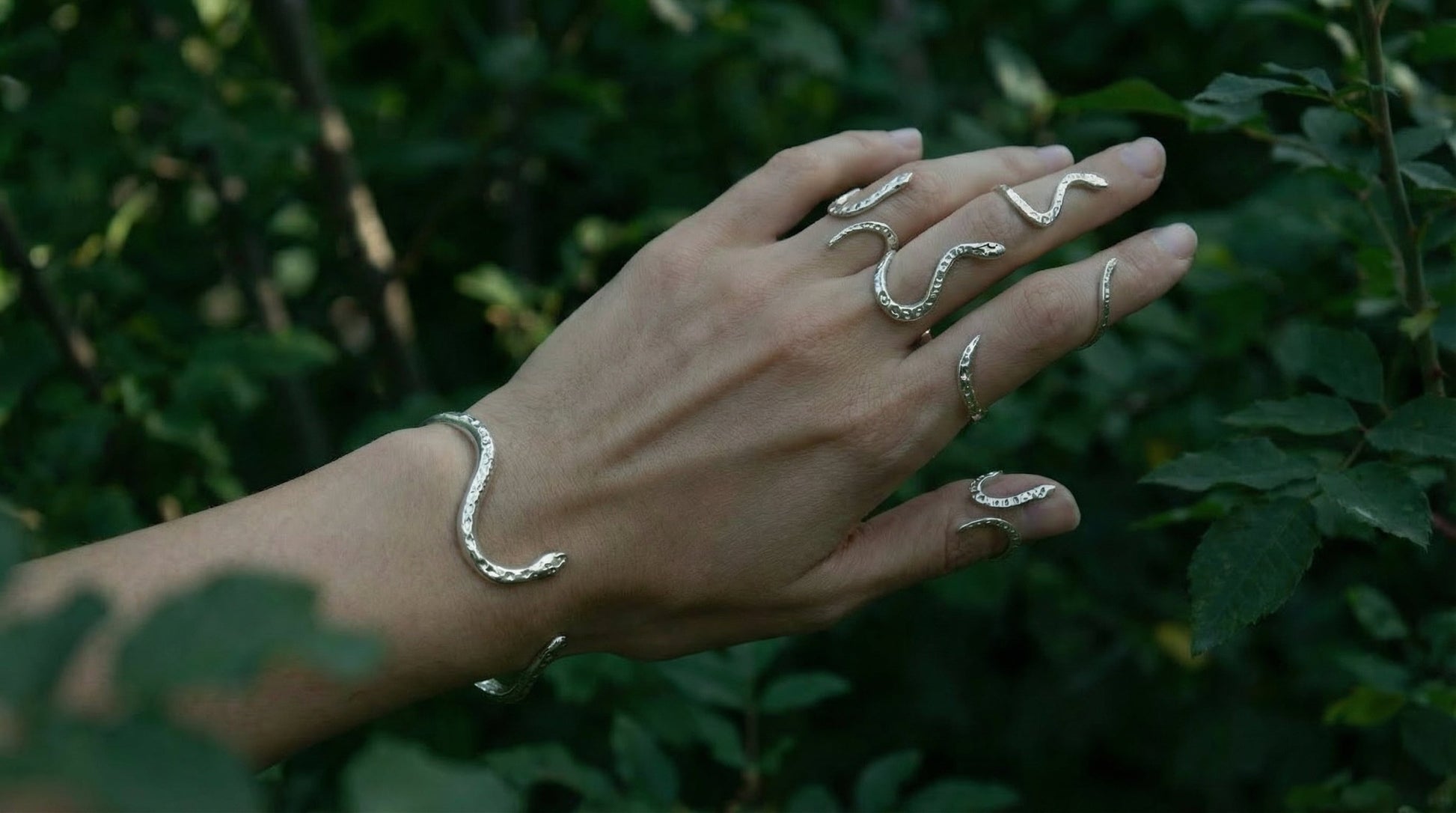 Hand adorned with sterling silver snake rings and bracelet, photographed in natural light among green foliage, designed in Italy.