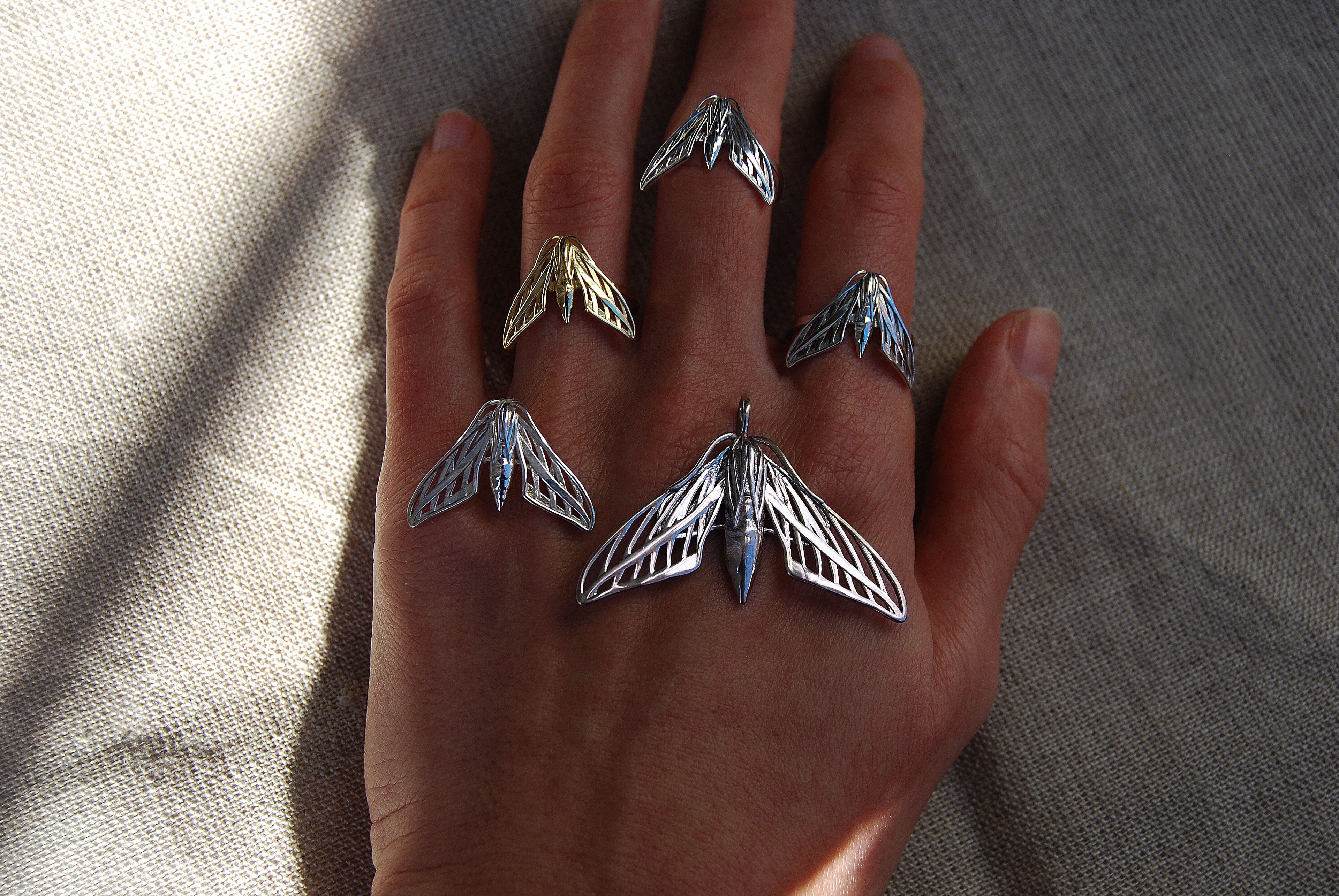 Full sphinx moth jewelry collection in silver and gold finishes worn on hand, featuring rings and matching pendant.