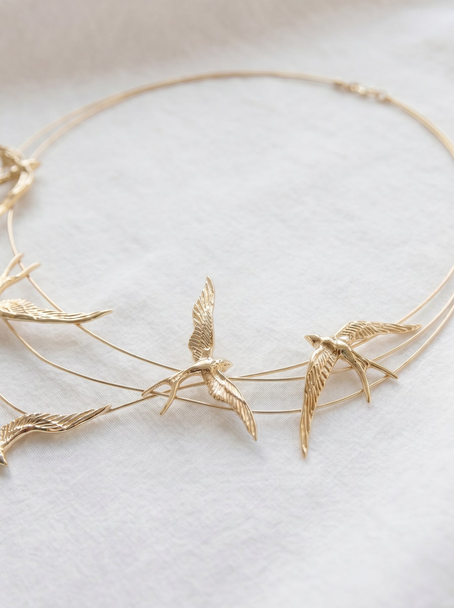 Close‑up of gold necklace with sculpted swallows in flight along fine arcs, softly lit on white fabric.