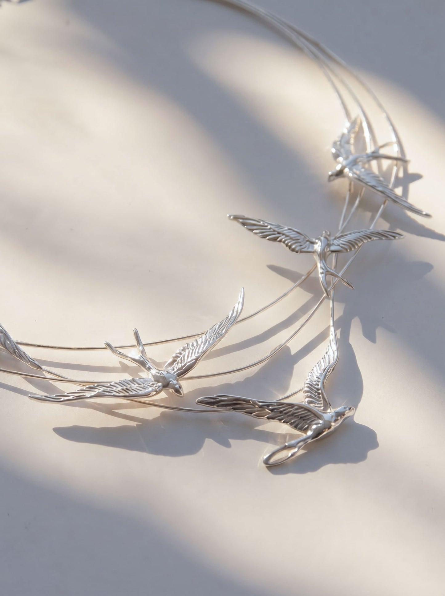 Swallow Arch Necklace photographed flat in minimal composition, showing the full arc of sculpted swallows and soft light reflections.