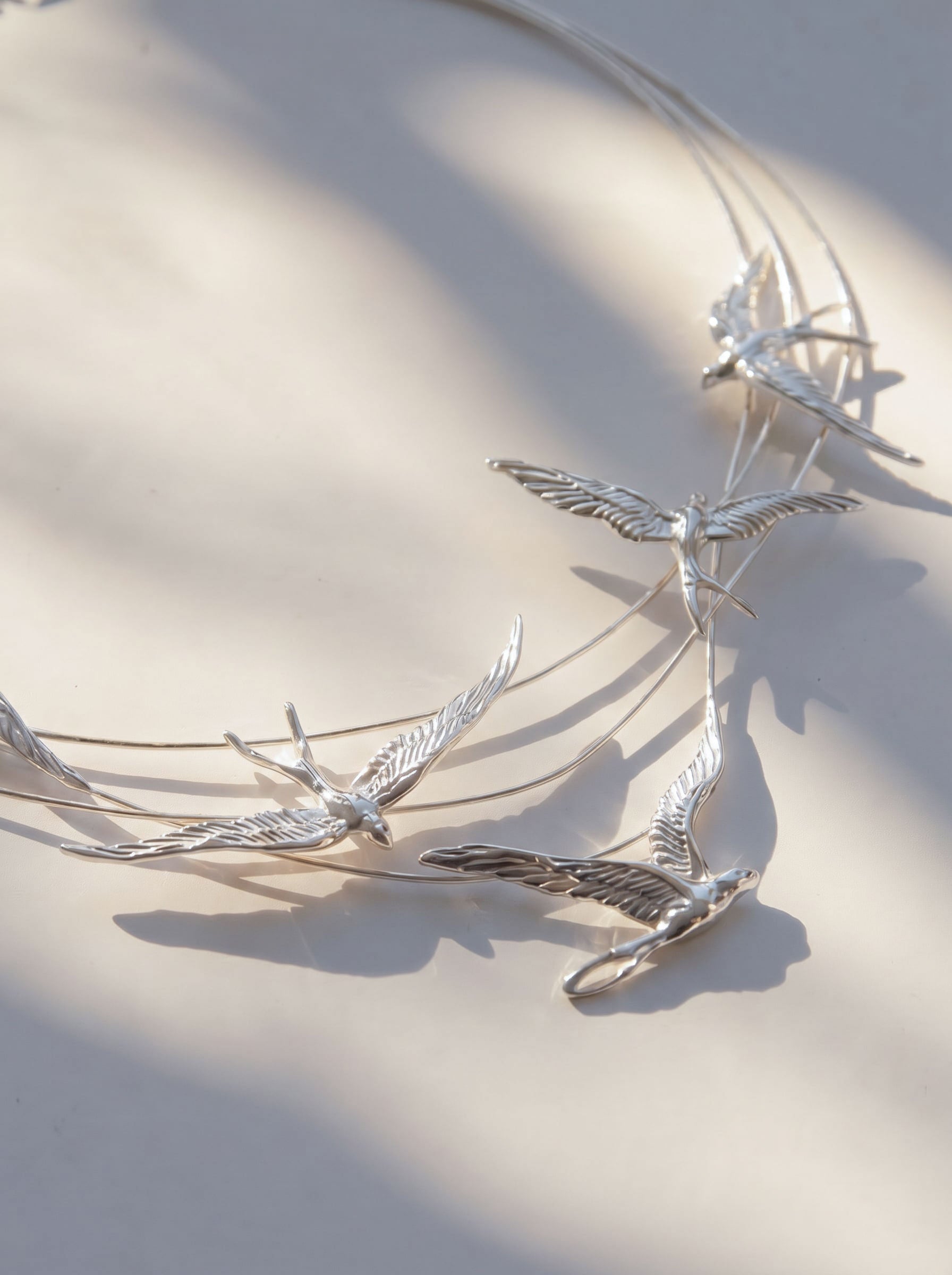 Swallow Arch Necklace photographed flat in minimal composition, showing the full arc of sculpted swallows and soft light reflections.