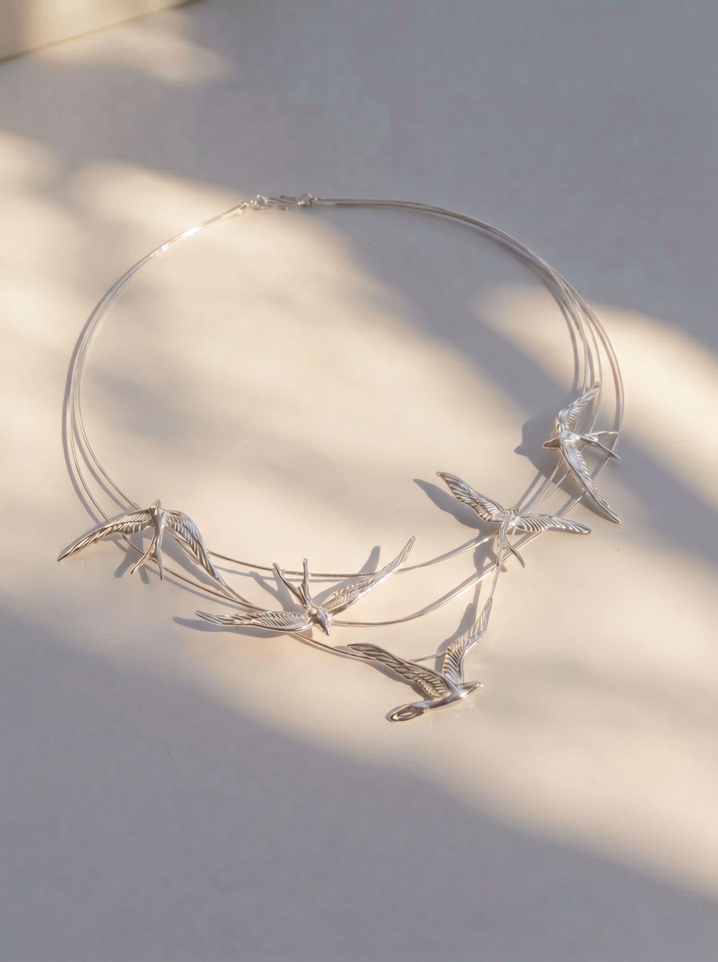 Swallow Arch Necklace photographed flat in sunlight, highlighting engraved feather textures and fluid silver curvature.
