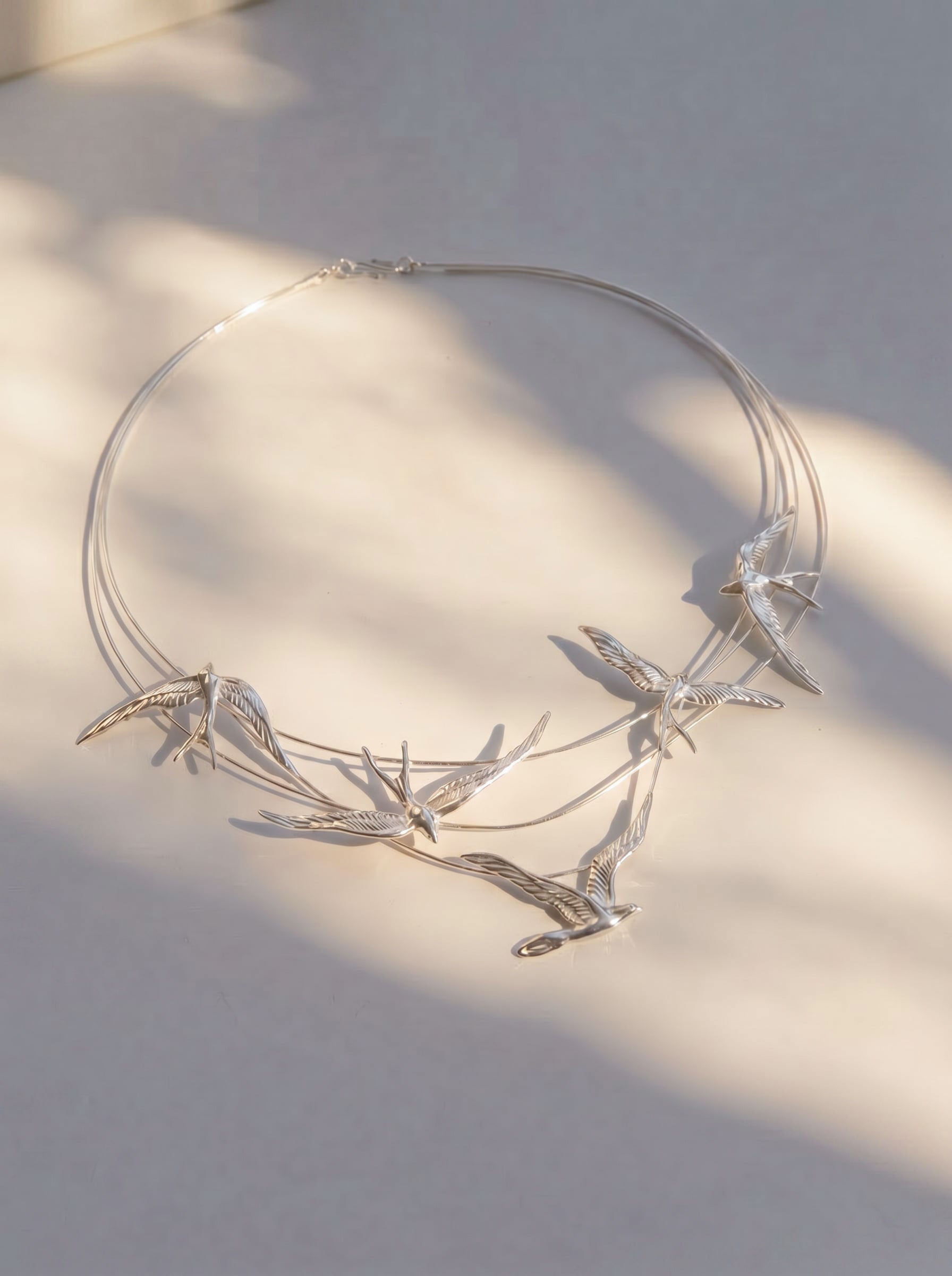 Swallow Arch Necklace photographed flat in sunlight, highlighting engraved feather textures and fluid silver curvature.