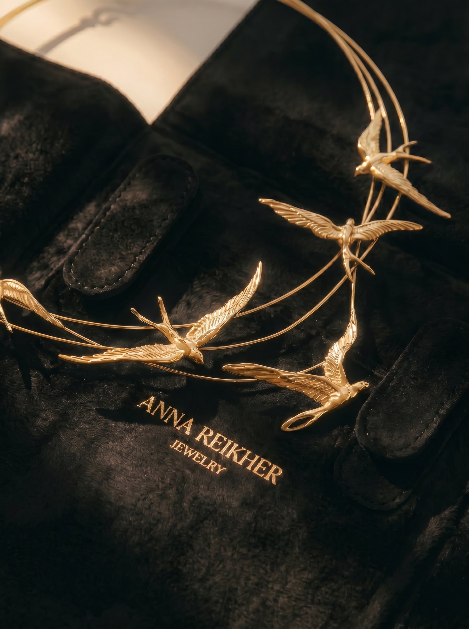 Close‑up of the Swallow Arch Necklace in gold, resting on black suede packaging embossed with “Anna Reikher Jewelry.” The sculptural swallows curve gracefully along fine arcs, catching warm light that highlights their wings and craftsmanship.