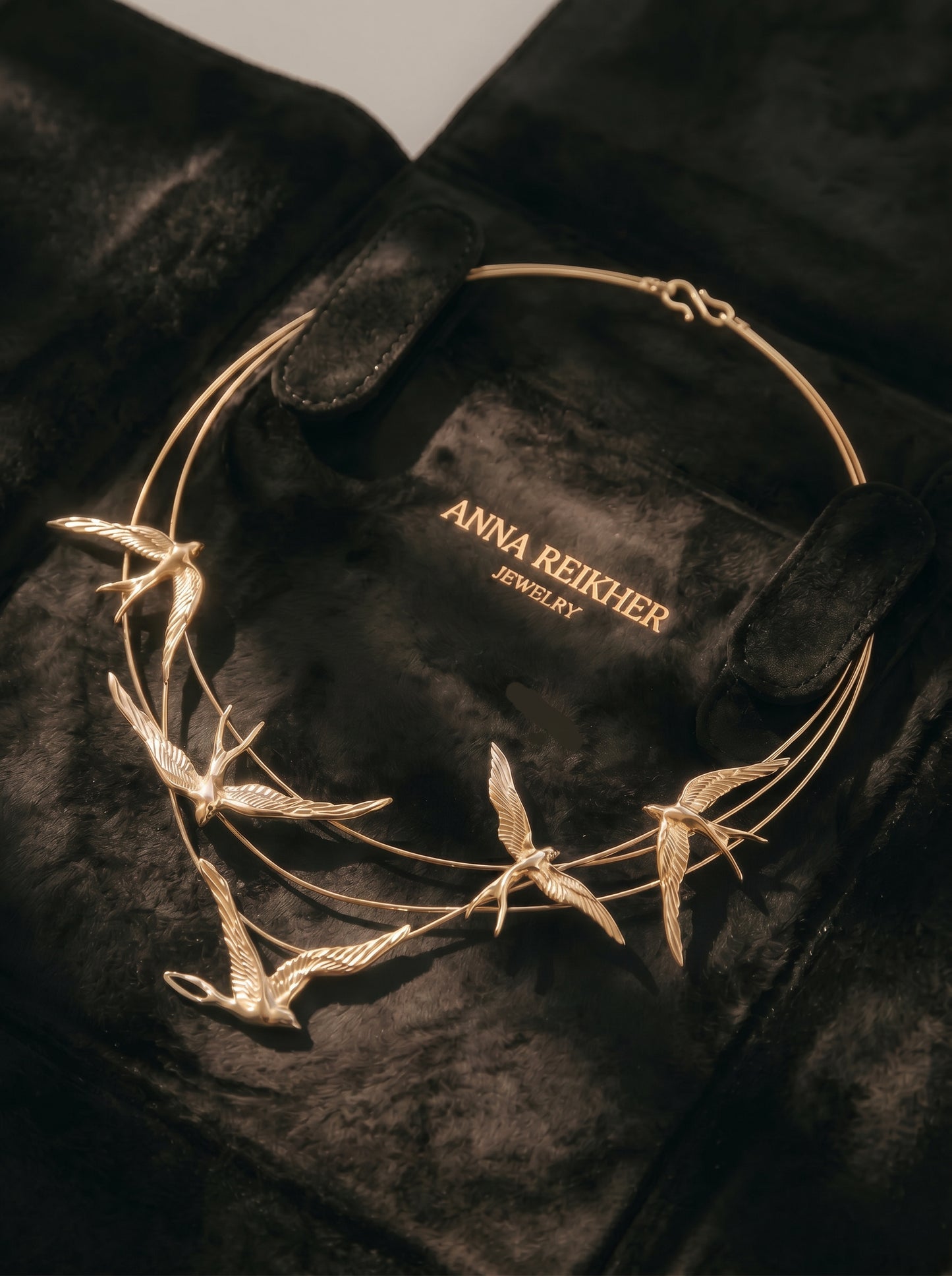 Gold Swallow Arch Necklace displayed on black suede packaging case embossed with “Anna Reikher Jewelry.” The sculptural swallows appear mid‑flight along fine arcs, glowing softly in warm light.