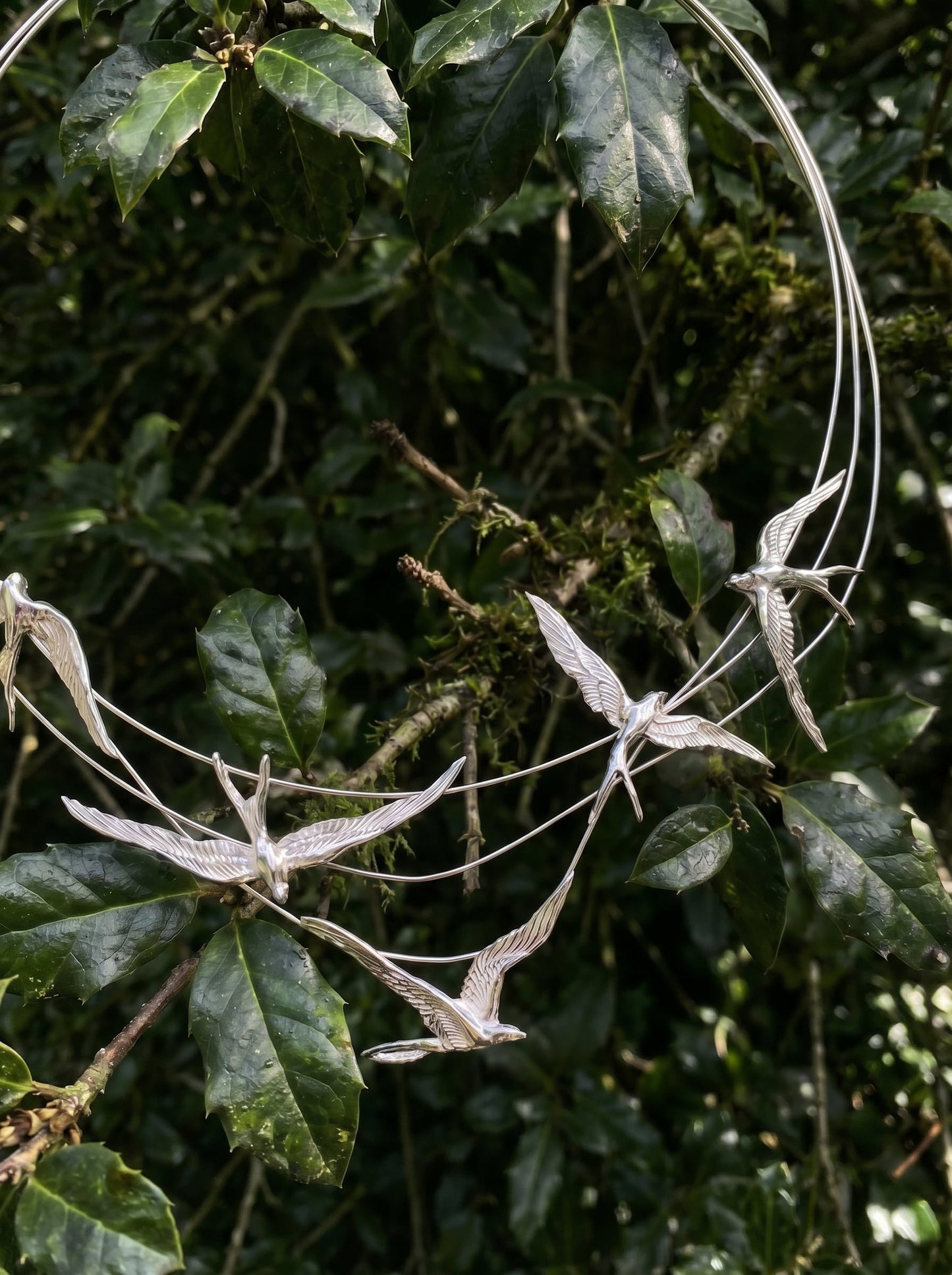 Close‑up of Swallow Arch Necklace among leaves, showing detailed bird figures and reflective silver surfaces.