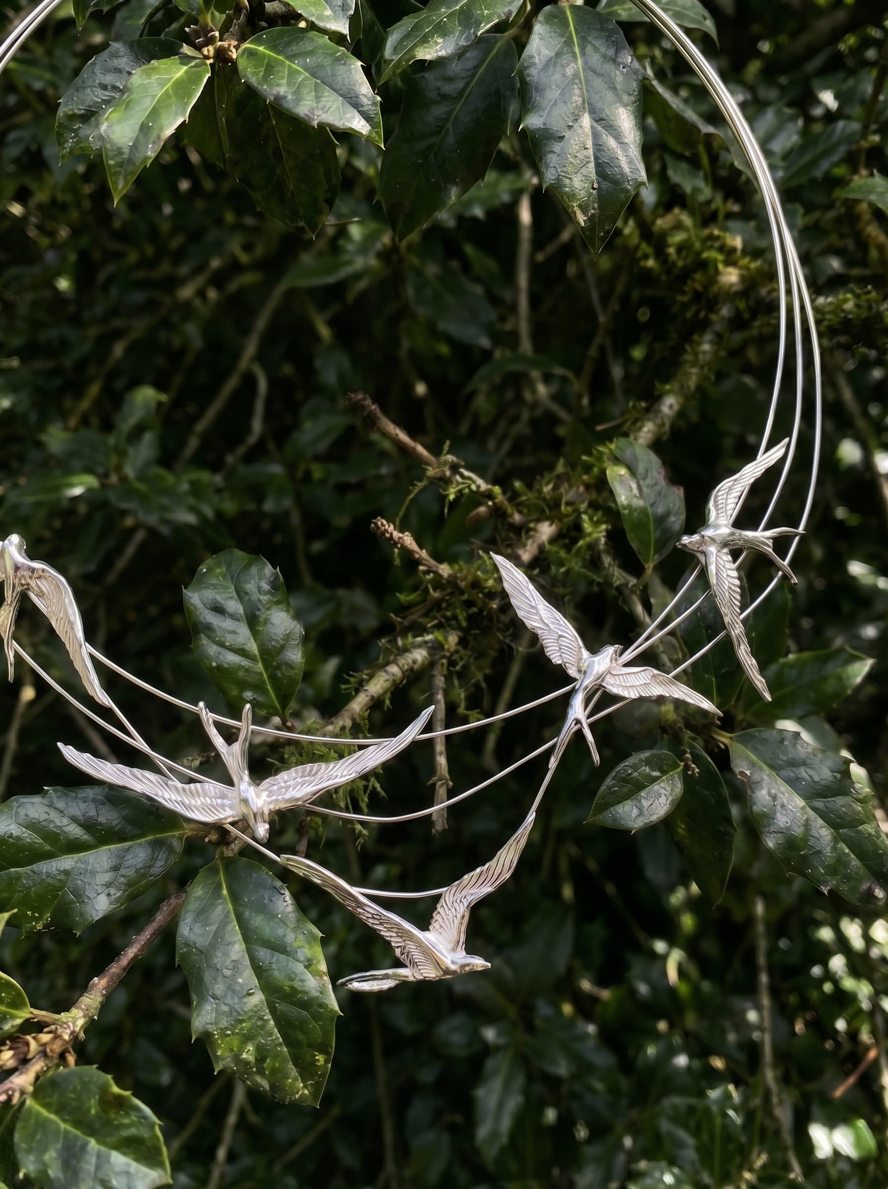 Close‑up of Swallow Arch Necklace among leaves, showing detailed bird figures and reflective silver surfaces.