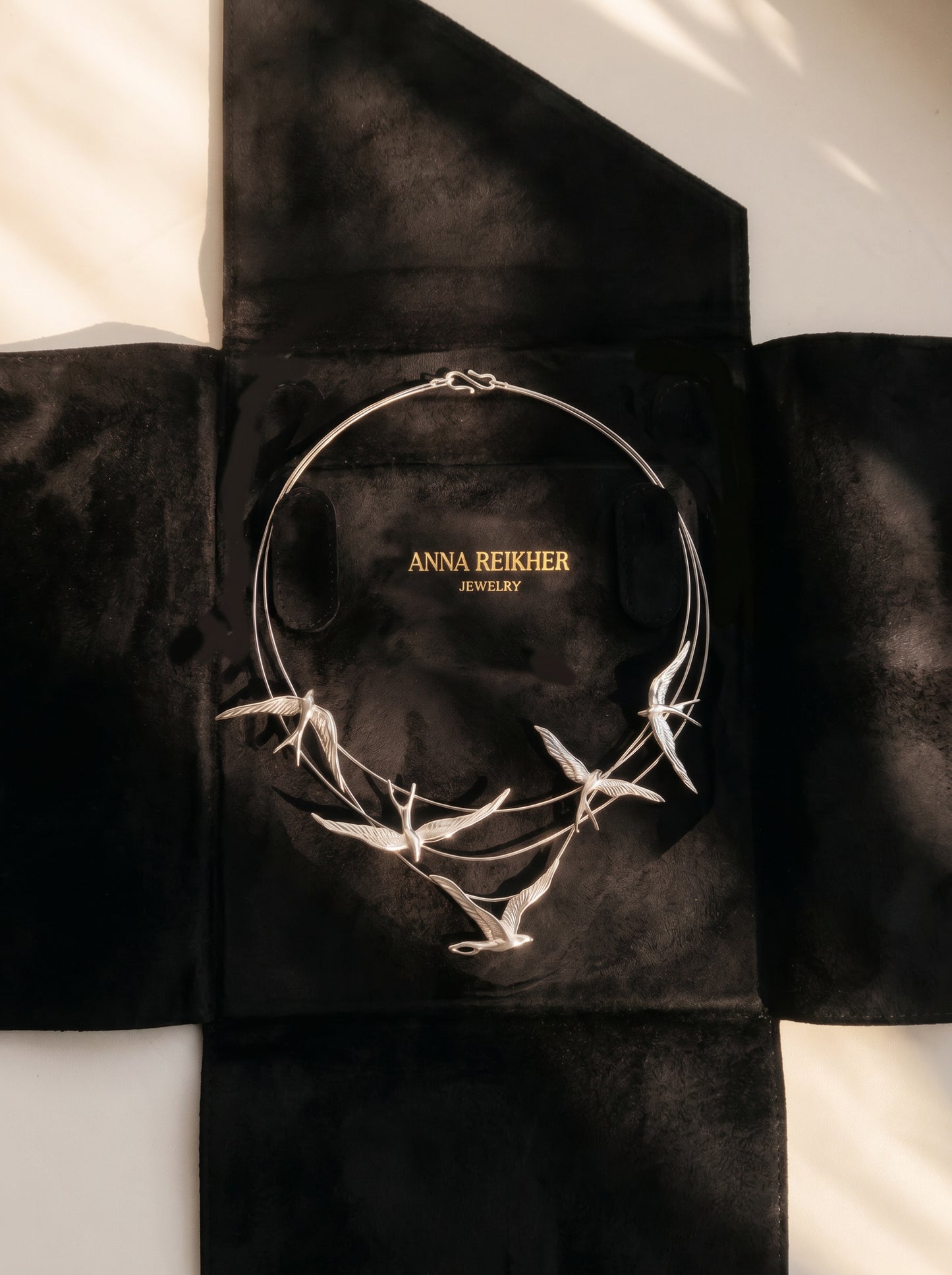 Silver Swallow Arch Necklace displayed on black suede packaging case embossed with “Anna Reikher Jewelry.” The sculptural swallows curve gracefully along fine arcs, catching soft light that highlights their wings and metallic sheen.