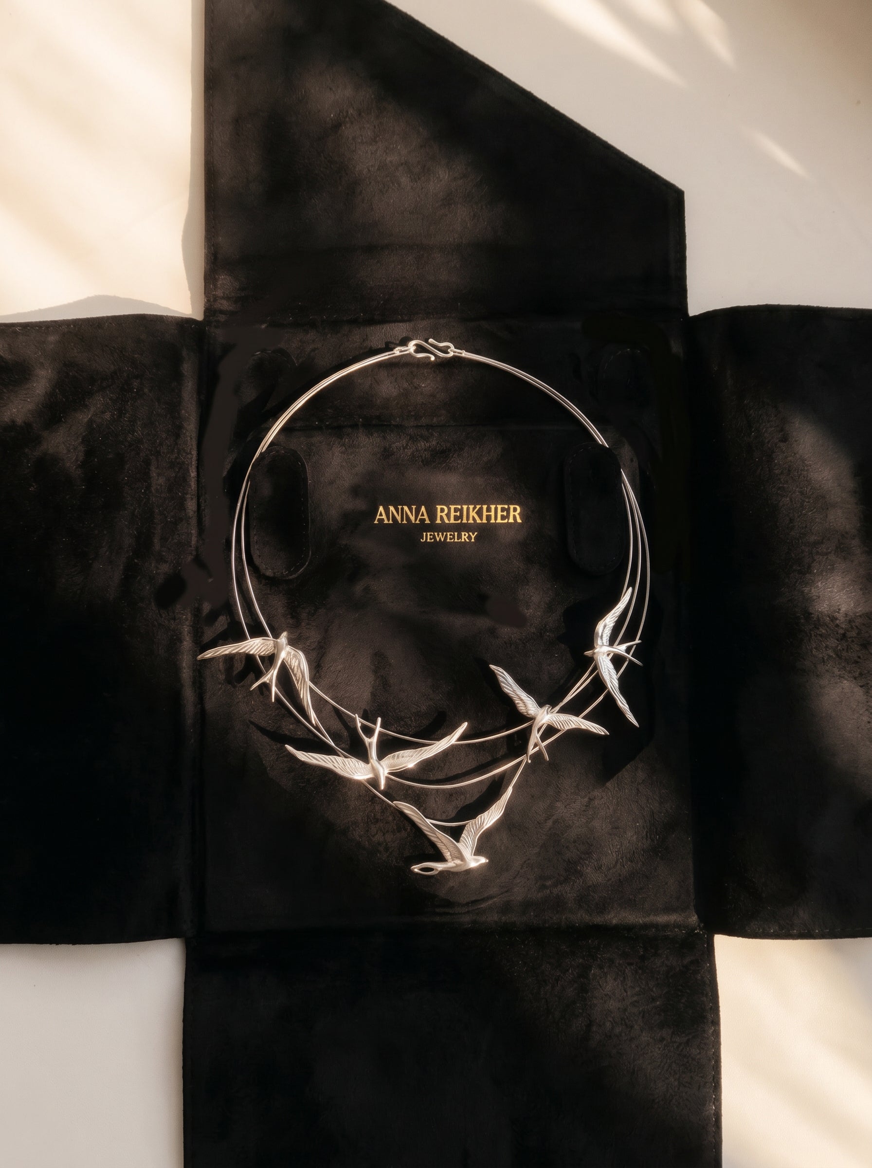 Silver Swallow Arch Necklace displayed on black suede packaging case embossed with “Anna Reikher Jewelry.” The sculptural swallows curve gracefully along fine arcs, catching soft light that highlights their wings and metallic sheen.