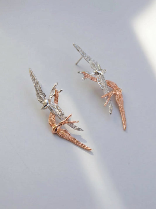Swallow Flight Stud Earrings in sterling silver and rose gold, photographed in sunlight, showing two sculpted birds mid‑flight with delicate wing engravings.