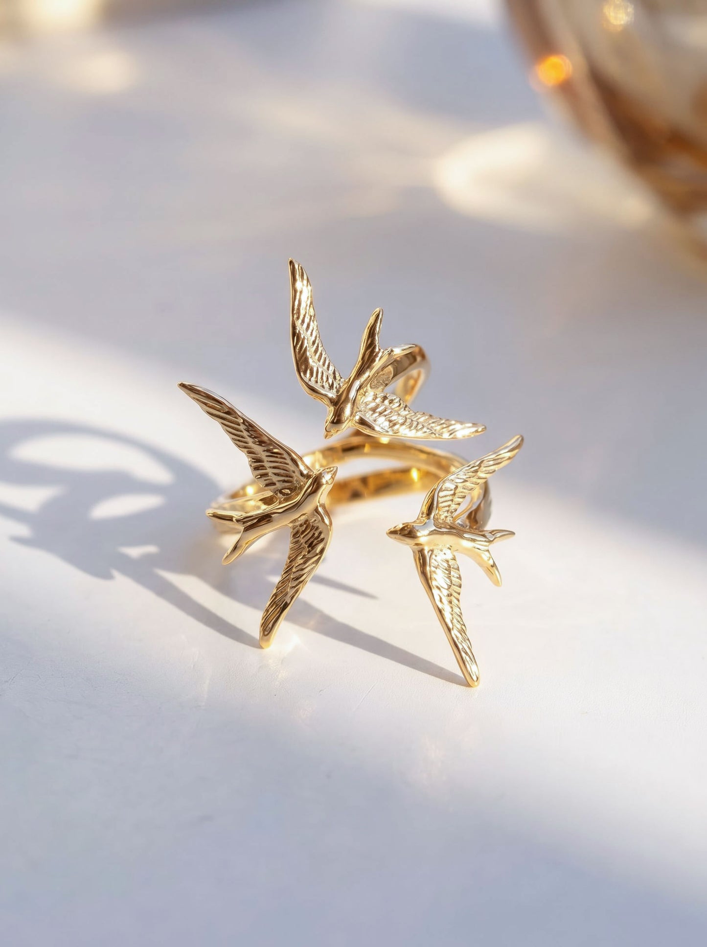 Swallow Gathering Ring in gold, photographed in sunlight, showing three sculpted birds curving inward toward a shared center.