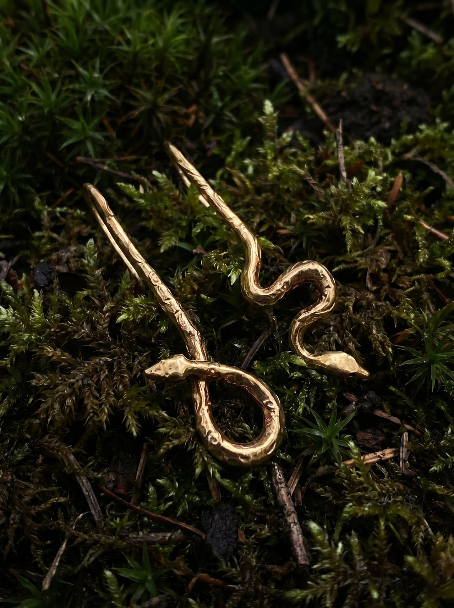 Pair of handcrafted gold serpent earrings with engraved texture, displayed on natural moss background.