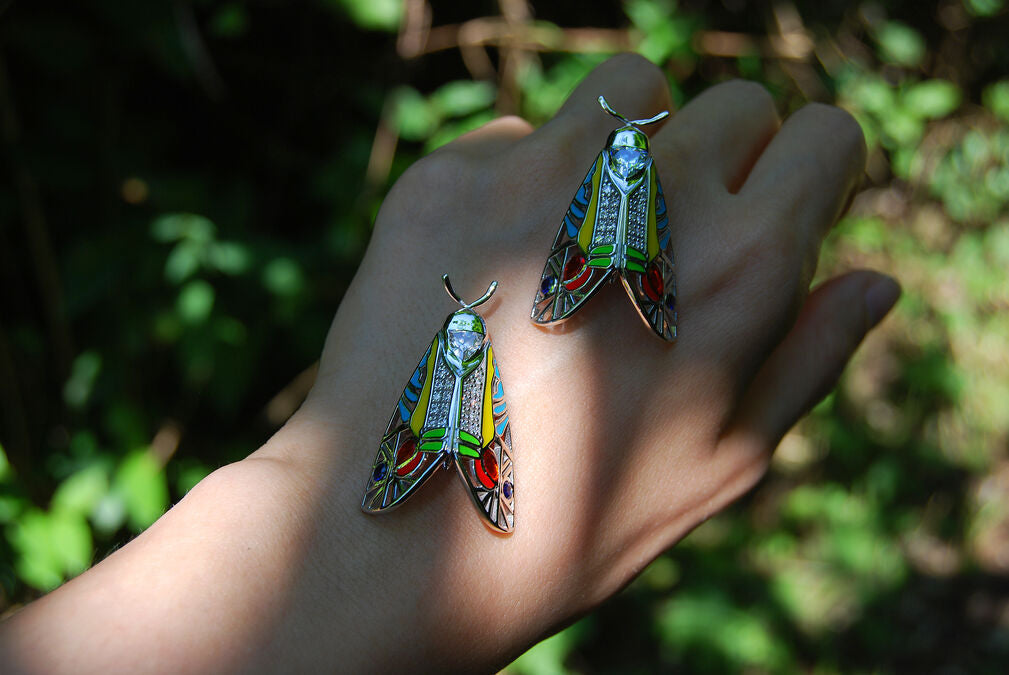 Two Picasso Moth pendants with colorful enamel and zirconia stones resting on hand against lush green forest background