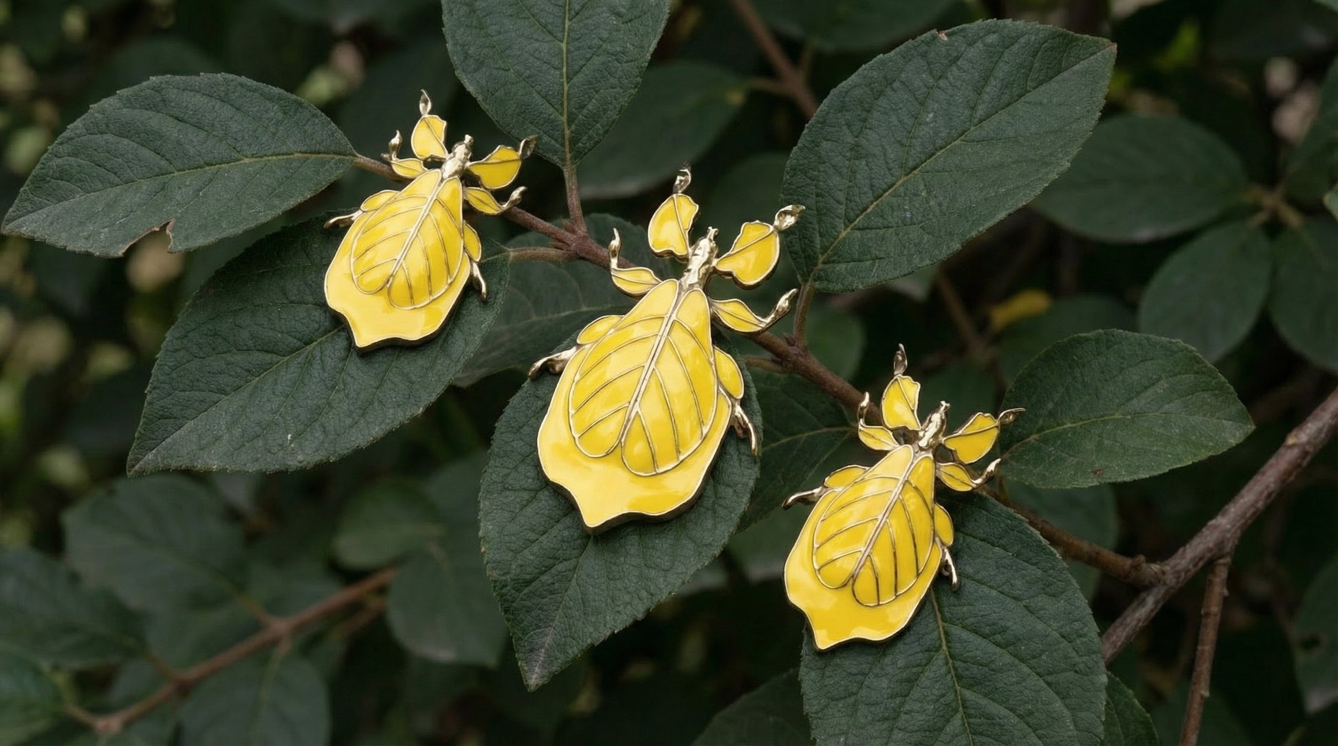 Three yellow enamel leaf‑insect pieces with gold outlines—two earrings and one pendant—resting on deep green leaves