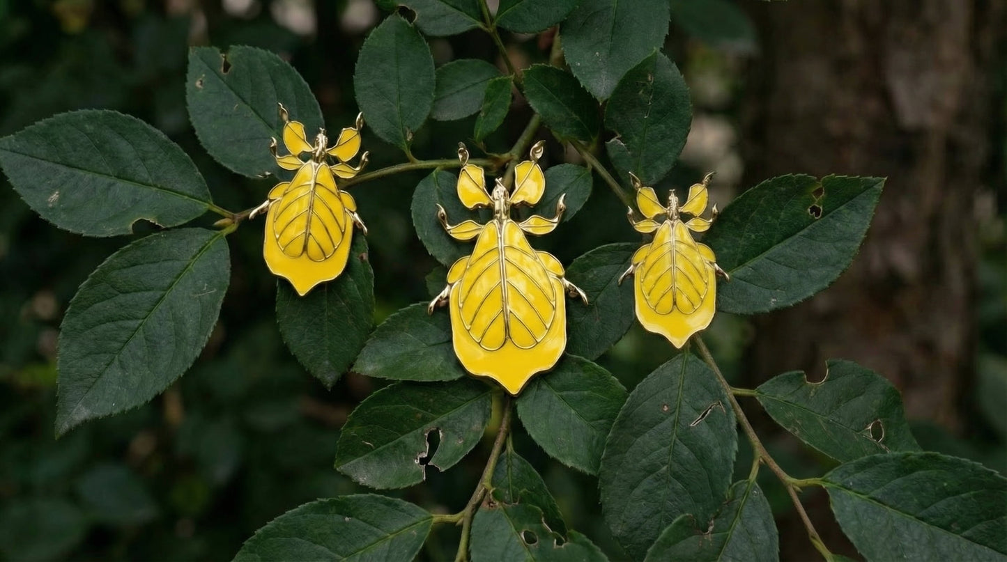 Three yellow leaf‑insect enamel pieces with gold edges resting on green leaves, showing their veined, leaf‑mimicking patterns.