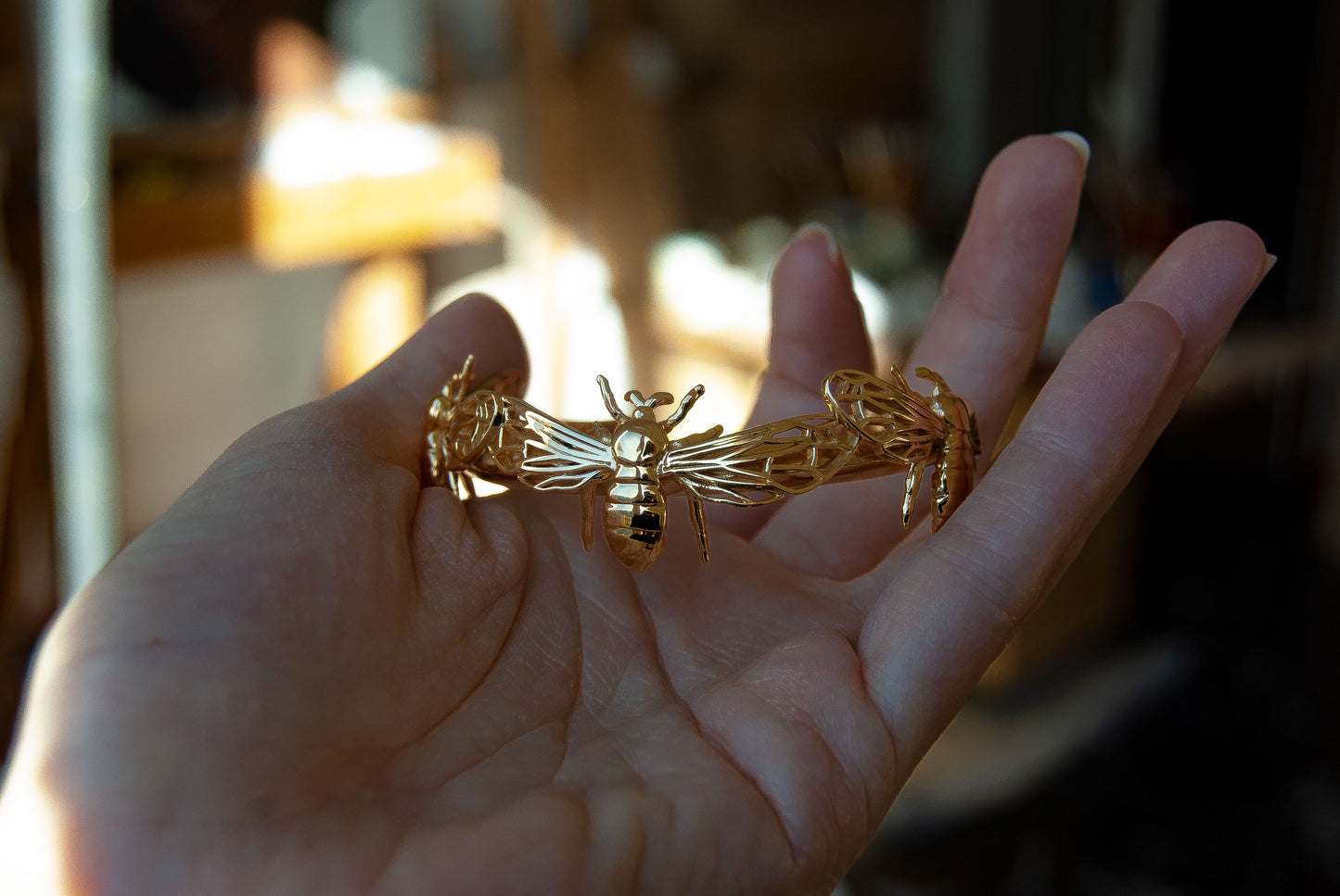 Hand holding gold bee bracelet cuff from front angle, showing three sculpted bees.