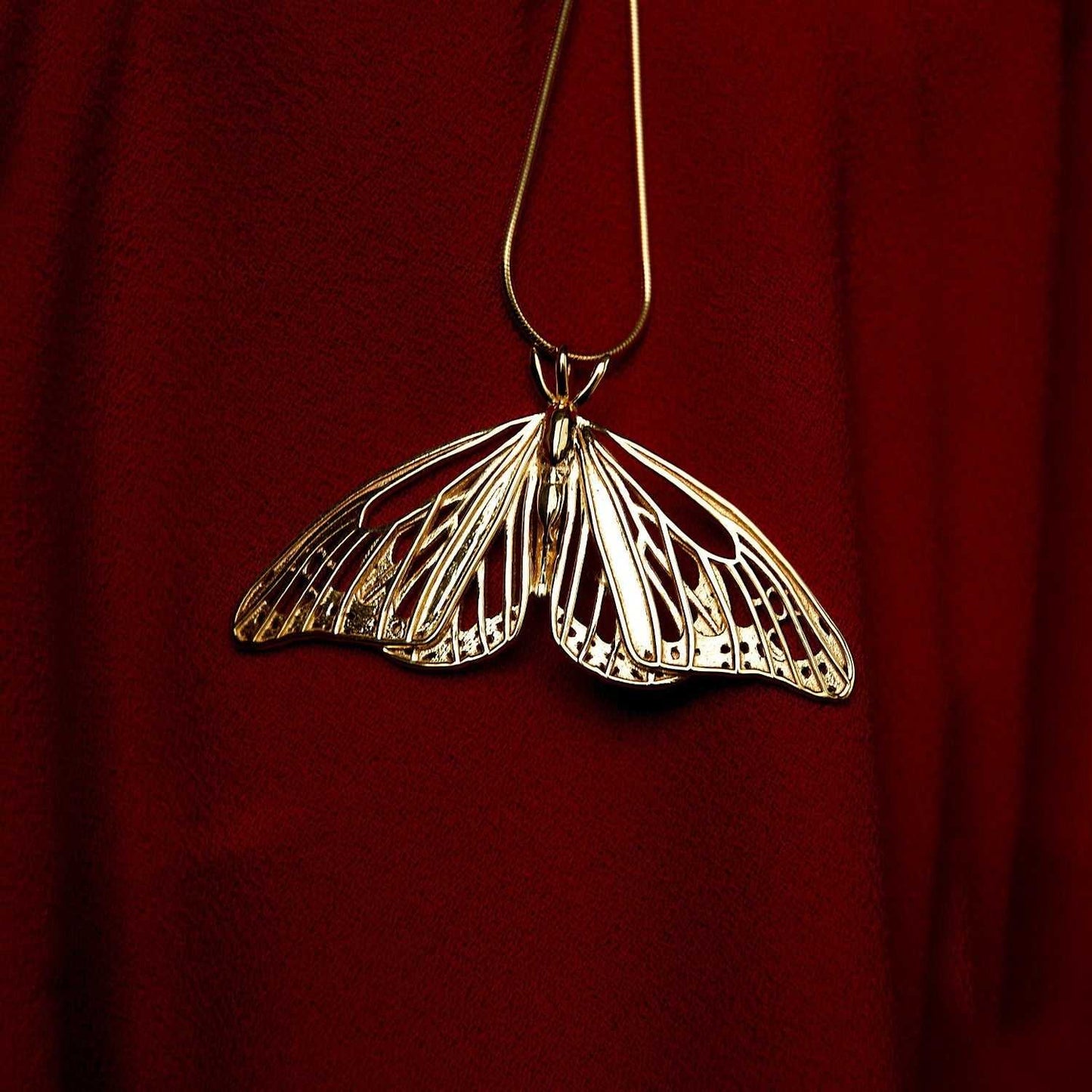 Gold Monarch Butterfly pendant displayed on deep red fabric, emphasizing its luminous finish.