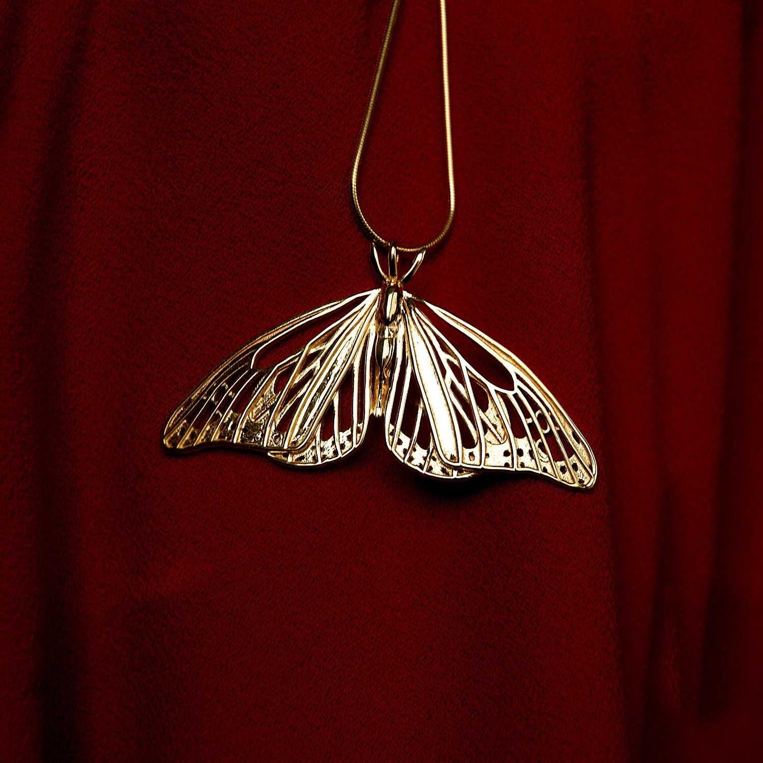 Gold Monarch Butterfly pendant displayed on deep red fabric, emphasizing its luminous finish.